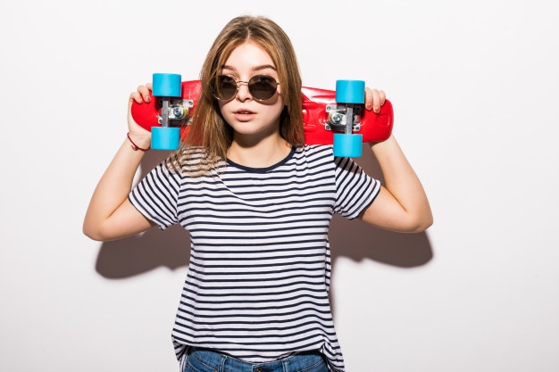 Portrait of young teen girl in sunglasses posing with skateboard while standing over white wall